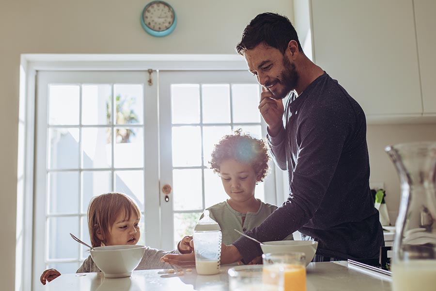 Contact Us - Smiling, Busy Dad Makes a Call While Fixing His Two Kids' Breakfast in Their Sunny Kitchen